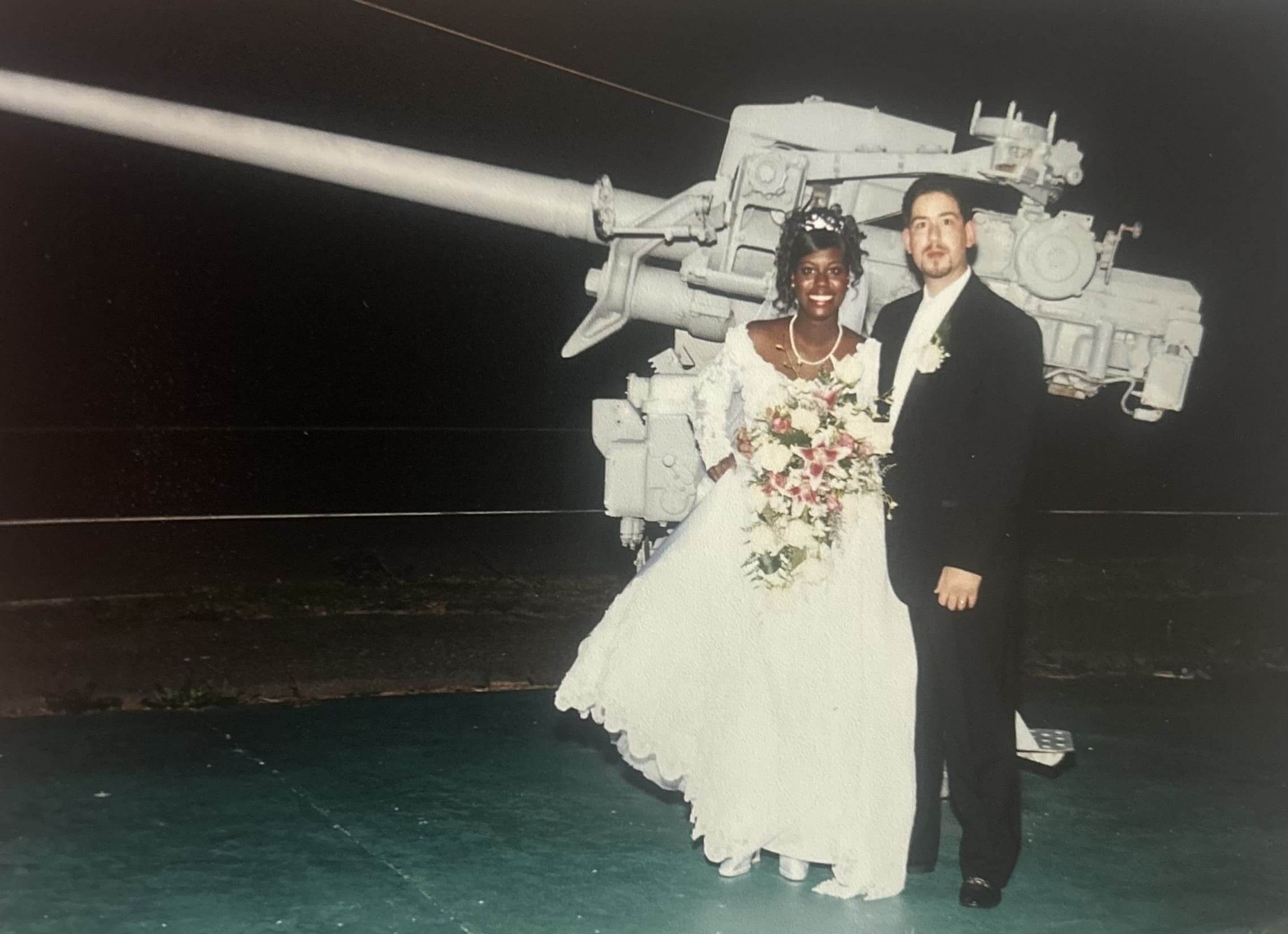 A bride and groom posing in front of a large artillery gun at night.