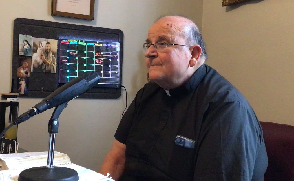 Priest sitting near a microphone, office background.